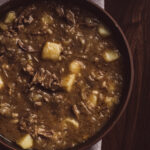 Austrian Goulasch flatlay image. The goulasch is in a brown bowl that is on top of a dark brown wooden surface.